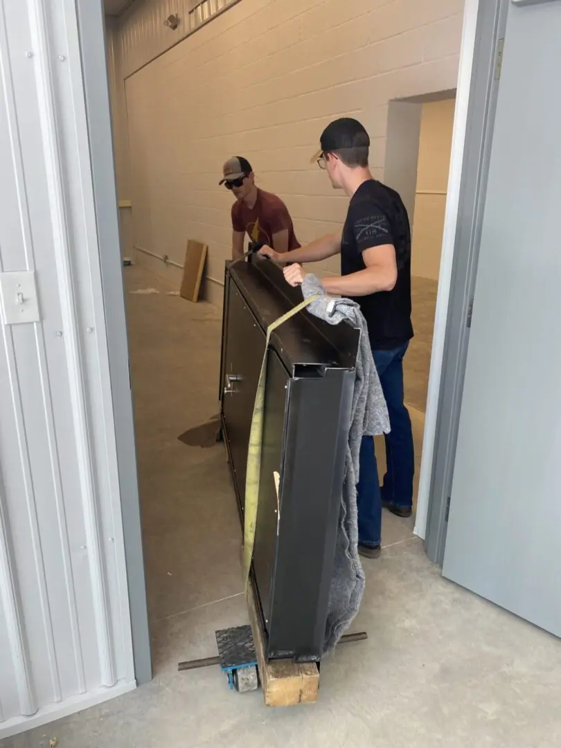 Two men moving a piece of luggage into a room.